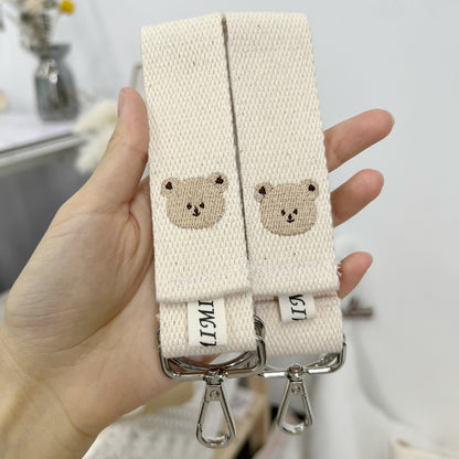 Two beige straps with bear face designs held by a hand against a blurred background.